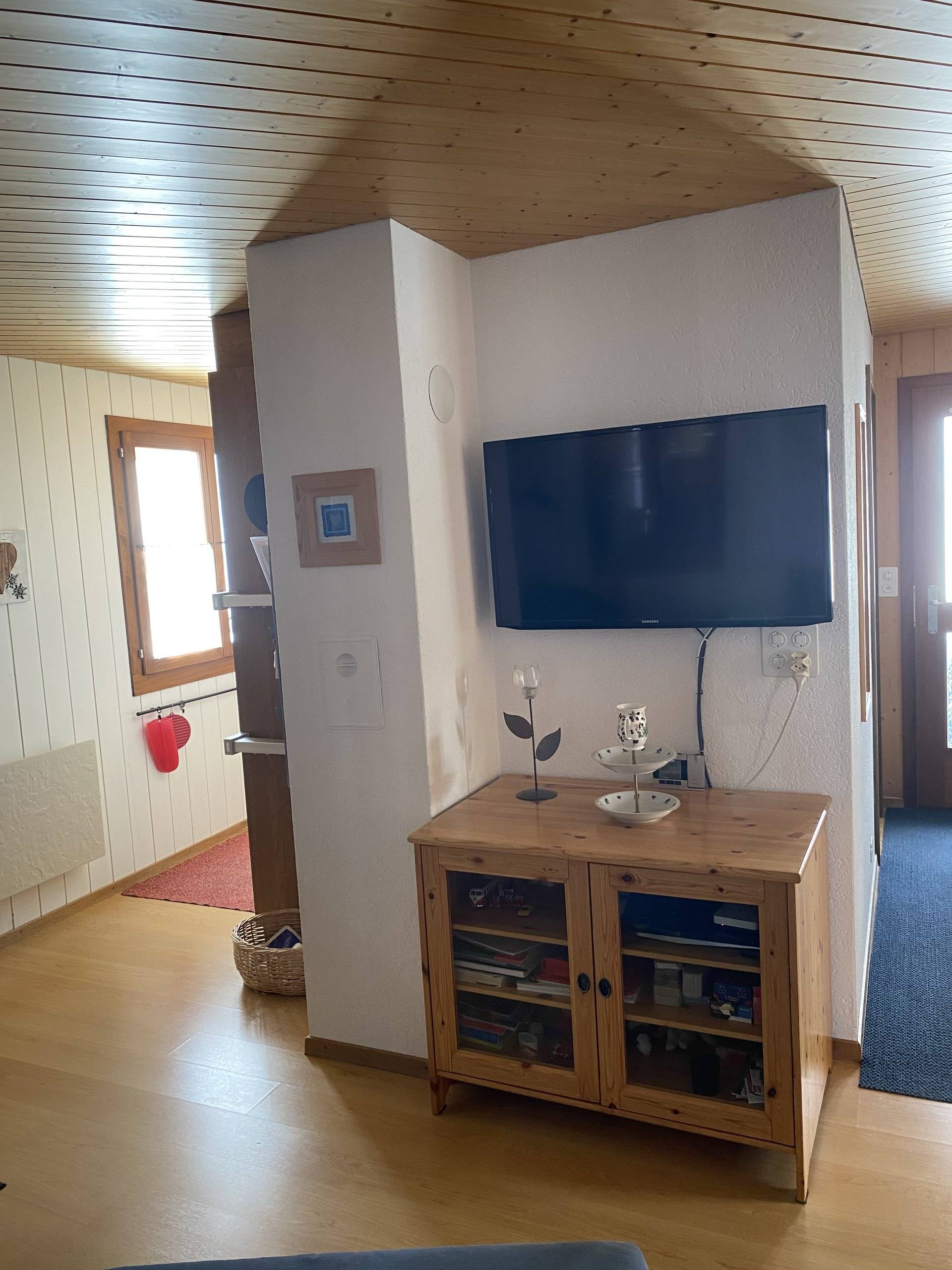 Eingangsbereich mit Holzkommode und Fernseher an der Wand, rechts der Weg ins Wohnzimmer