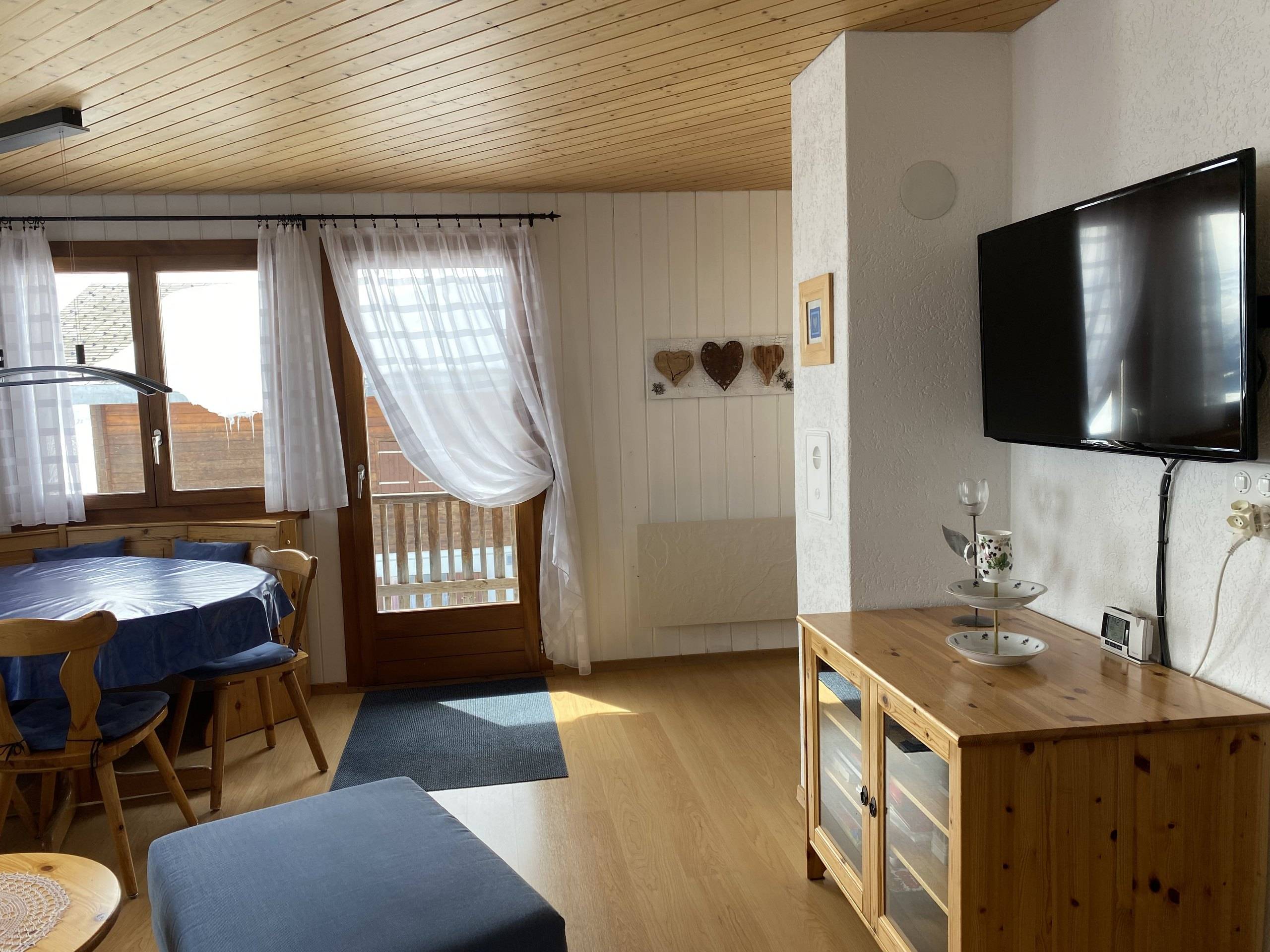 Wohnzimmer mit Essplatz, Balkontür, Fernseher an der Wand und Holzkommode