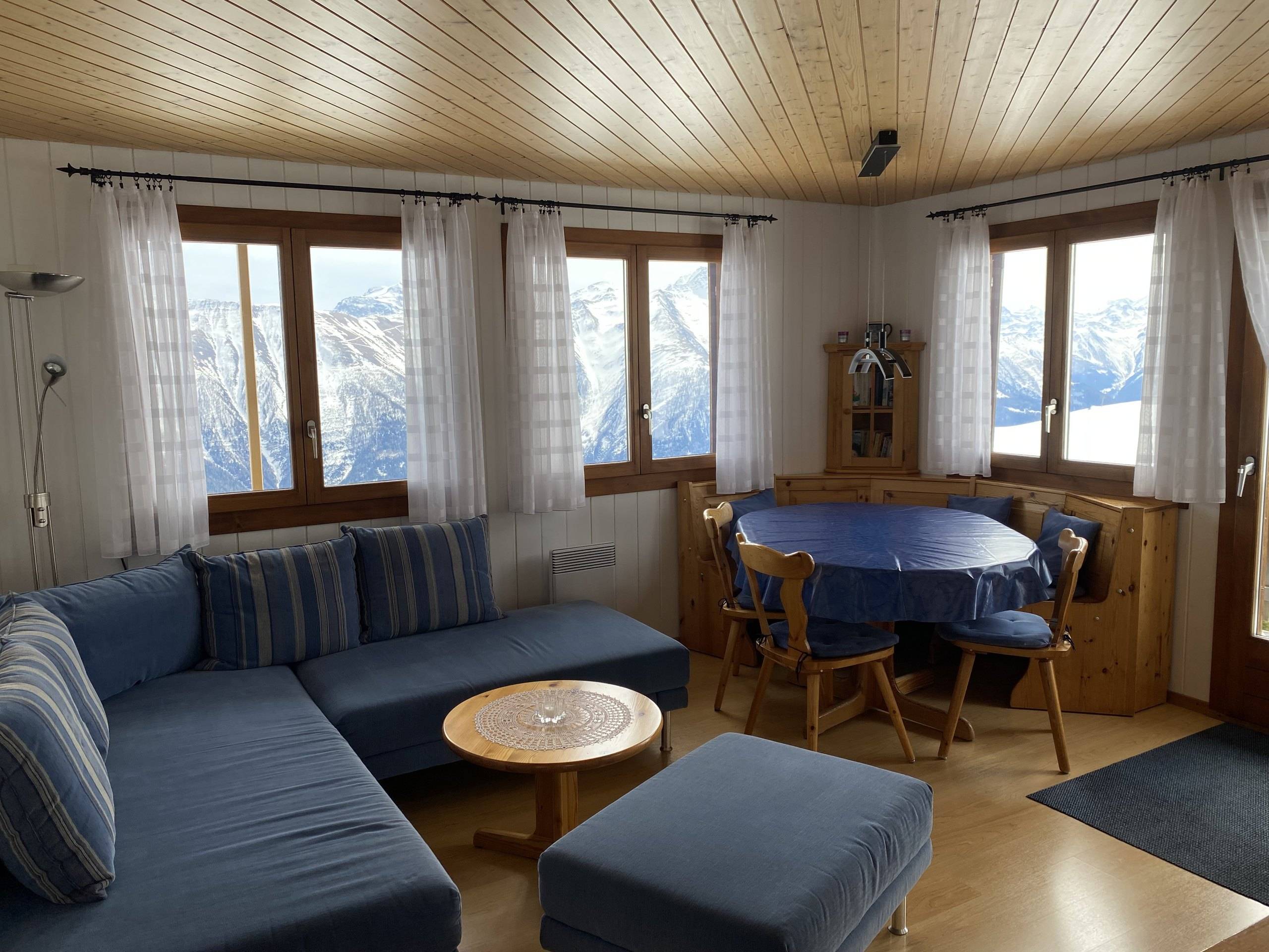 Wohnzimmer mit blauem Ecksofa, Salontisch und Essplatz vor zwei Fenstern mit Bergblick